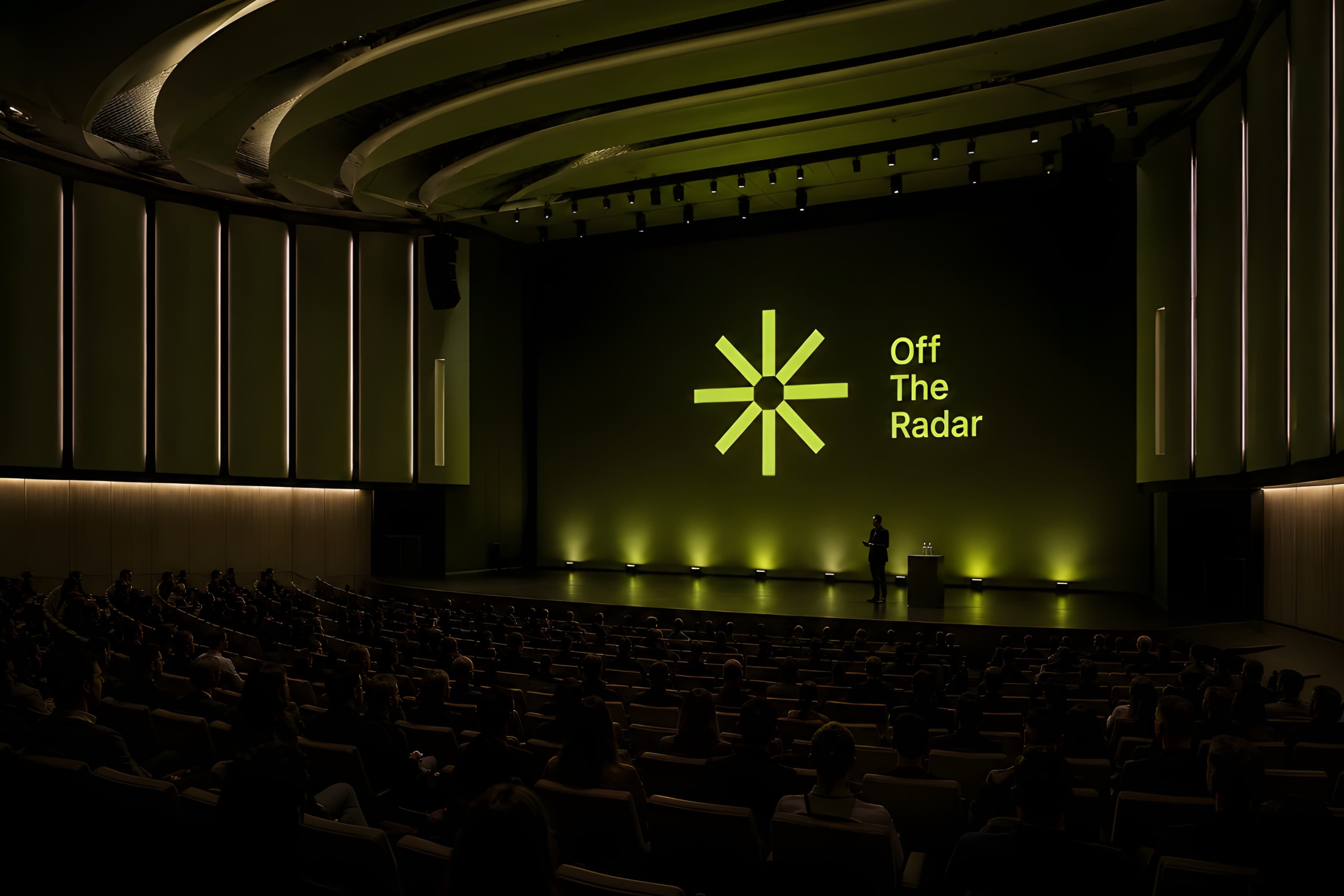 A speaker on stage at Off the Radar, in front of a green-lit Off the Radar wordmark and 8-spoke asterisk, with a full auditorium watching.